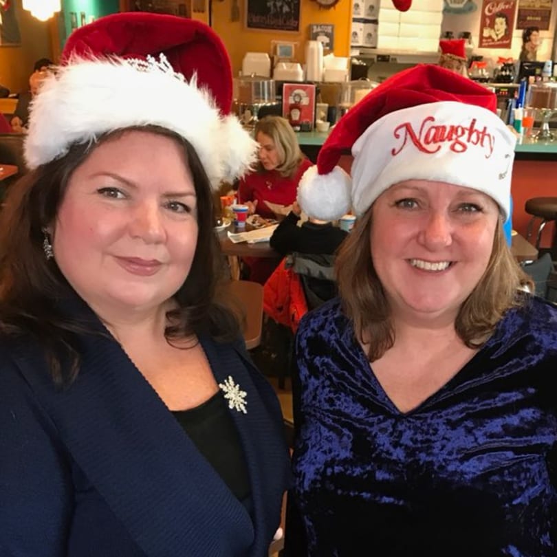 Two women in festive Santa hats, one with "Naughty" embroidered, smiling in a diner.