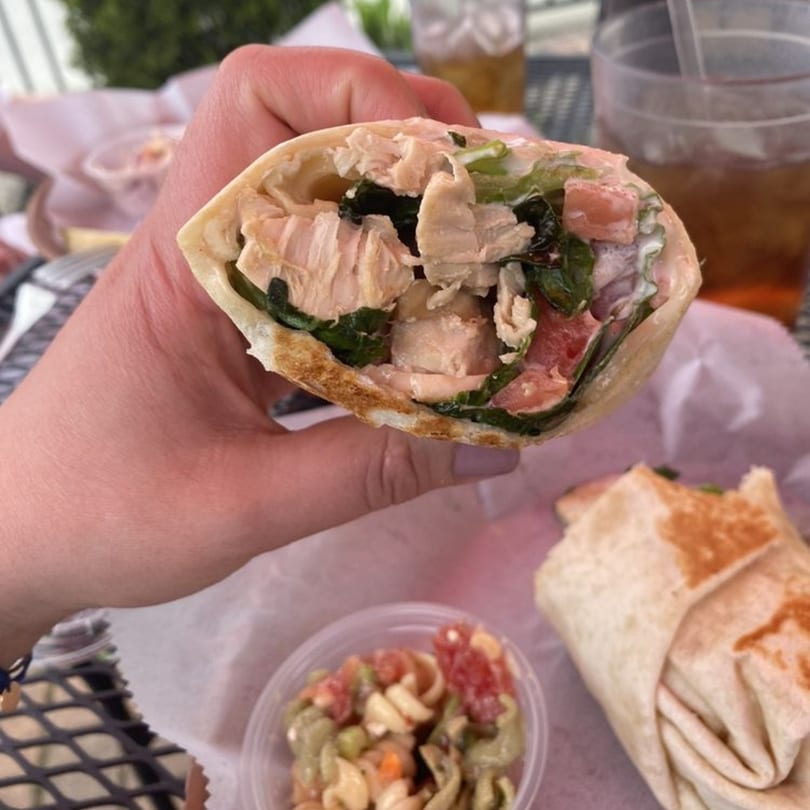 A hand holds a chicken wrap filled with spinach and tomatoes, with pasta salad and iced tea nearby.