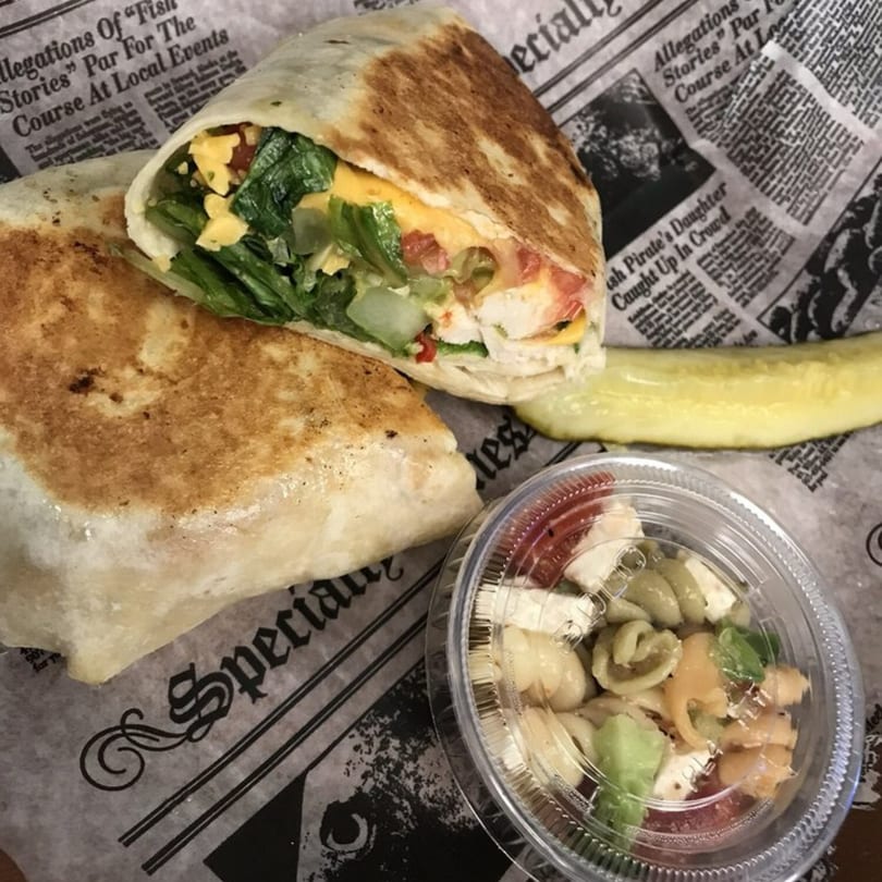 A grilled chicken wrap with lettuce, cheese, and tomatoes, served with pasta salad and a pickle.