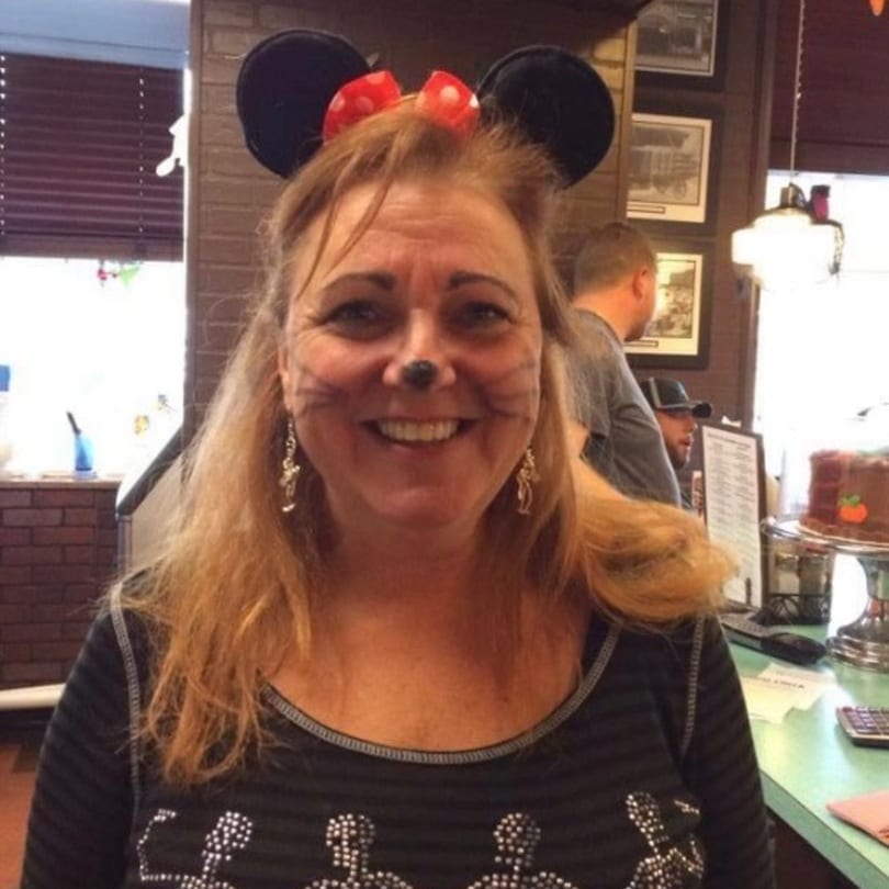A smiling woman with Minnie Mouse ears and painted whiskers stands in a diner.