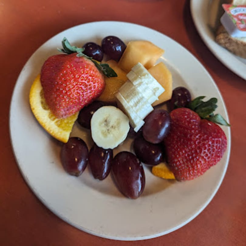 A white plate of fresh fruit, including strawberries, grapes, banana slices, cantaloupe, and orange.